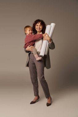 multitasking woman, professional achievements, time management, happy mother holding in arms daughter and rolled paper blueprints on grey background, building successful career, full length  clipart