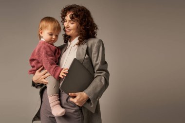 profesyonel emeller, modern ebeveynlik, iş hayatı uyumu, neşeli çalışan anne dizüstü bilgisayarı ve kızını gri arka planda tutmak, zaman yönetimi, kendine güven 