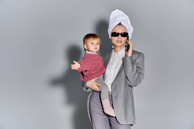 multitasking, woman in sunglasses holding in arms toddler daughter and standing with towel on head, balancing lifestyle, businesswoman in formal wear talking on smartphone on grey background  clipart