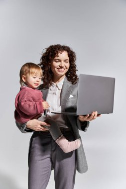 time management, happy working mother holding laptop and toddler daughter in arms on grey background, work life harmony, career and family, modern parenting, professional success, businesswoman  clipart