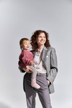 career and family, happy working mother holding toddler daughter in arms and standing with hand in pocket on grey background, work life balance, modern parenting, professional success, businesswoman  clipart