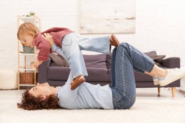 Kaliteli aile zamanı, mutlu anne küçük kızını kaldırıyor ve rahat oturma odasında halıya uzanıyor, iş hayatı dengesi, kot kıyafetler, günlük kıyafetler, aile ilişkileri, modern ebeveynlik 