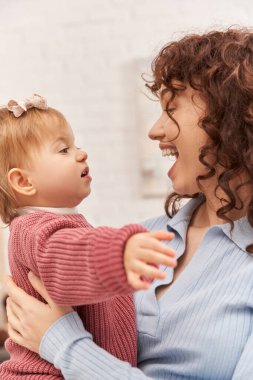 Dengeli bir yaşam tarzı, kıvırcık saçlı, çalışan bir anne kollarında kız bebek, hayrete düşmüş bir kadın ve küçük kızı, modern ebeveynlik, iş hayatı uyumu, bağ kurma ve çocukla yakınlaşma, mutluluk. 
