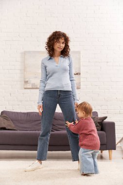 Kıvırcık annesinin, rahat oturma odasının yanında ilk adımlarını atan bir kız çocuğu, kot pantolonu, günlük kıyafetleri, aile zamanı, modern ebeveynlik, iş hayatı dengesi. 