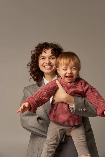 stock image quality time, career and family, smiling woman in suit holding daughter on grey background, career and family, loving motherhood, lifestyle, multitasking, work life balance concept 