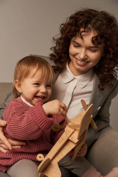 stock image successful mother, balancing work and life concept, happy businesswoman in suit sitting on armchair and playing with toddler daughter, wooden biplane, grey background, engaging with child 