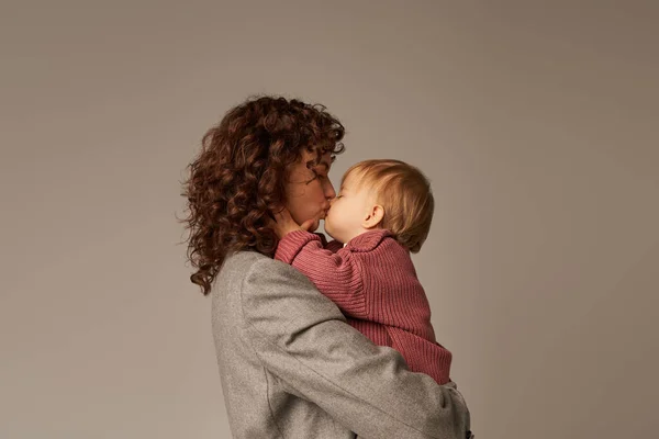 working mother, parenting and career, curly businesswoman holding in arms and kissing her toddler daughter on grey background, work life harmony concept, loving motherhood, quality time, side view