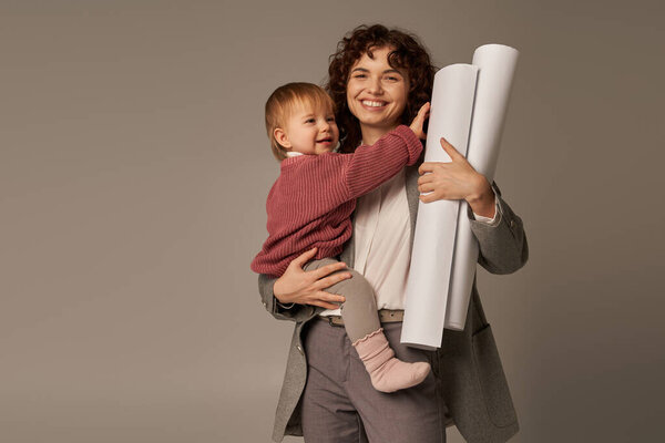 multitasking woman, professional achievements, time management, joyful mother holding in arms daughter and rolled paper blueprints on grey background, building successful career 