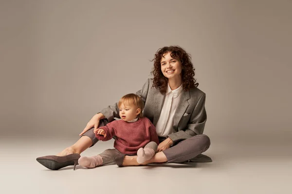 career and family, balancing between work and life, cheerful mother with curly hair hugging toddler daughter on grey background, quality time, modern parenting, businesswoman, full length