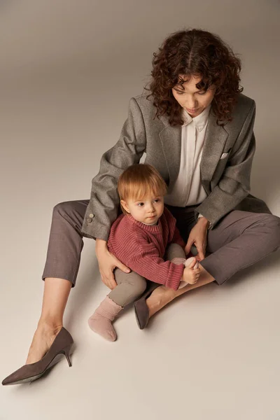 Stock image career and family, balancing between work and life, cheerful mother with curly hair hugging toddler daughter on grey background, quality time, modern parenting, businesswoman, high angle view 