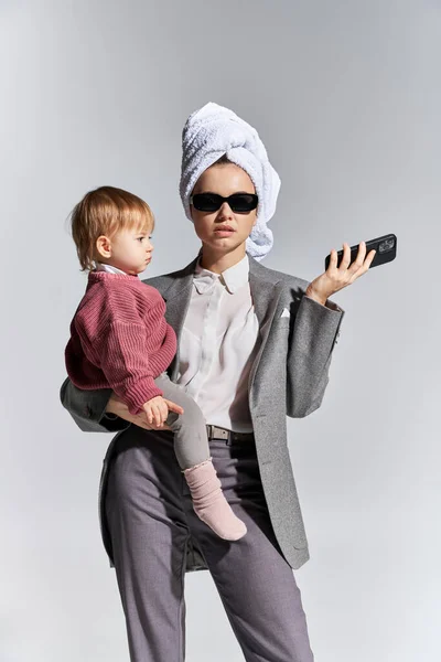 キャリアと家族マルチタスク腕の中で抱えているサングラスの女性幼児娘頭の上にタオルで立って灰色の背景にスマートフォンを保持している正式な摩耗のビジネス女性 — ストック写真