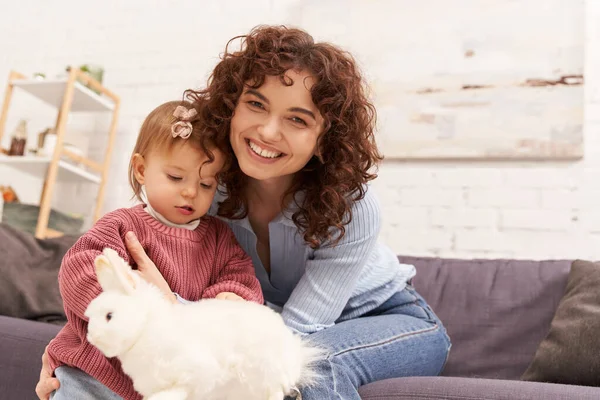work life balance, happy and curly woman sitting on couch with toddler daughter in cozy living room, playing with rabbit, quality family time, casual attire, bonding between mother and child