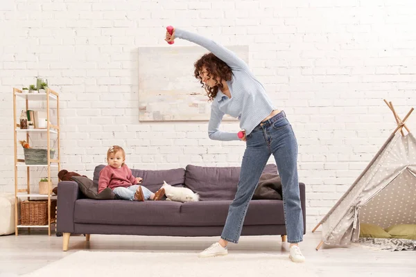 busy mom, modern parenting, working mother, balanced lifestyle, curly woman exercising with dumbbells near toddler daughter on couch in cozy living room, home workout, sport, time management