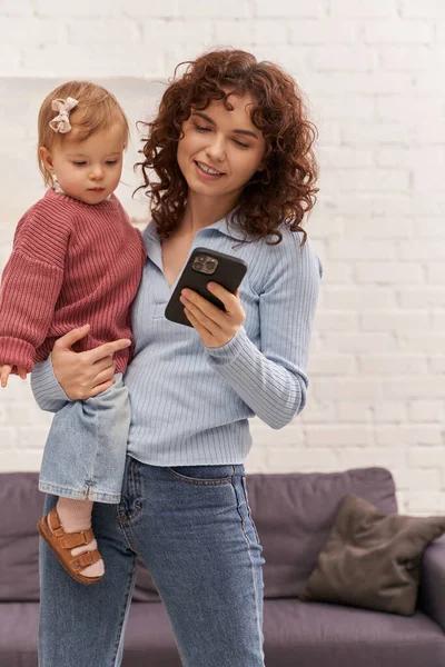 Encaracolado Jovem Segurando Braços Bebê Menina Usando Smartphone Equilíbrio Entre — Fotografia de Stock