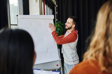 Genç ve sakallı tıp eğitmeni, ön planda bulanık katılımcıların bulunduğu eğitim odasındaki ilk yardım semineri sırasında ters yüz çizelgesini düzelterek acil durum müdahale kavramına katılıyor.