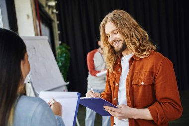stylish and long haired man smiling while writing on clipboard near woman and medical instructor with flip chart on blurred background, first aid seminar, health care and emergency response concept clipart