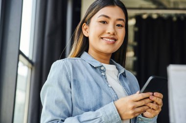 Genç ve modaya uygun Asyalı iş kadını esmer saçlı ve mutlu gülüşlü cep telefonuyla iletişim kuran çağdaş ofis kameralarına bakan hırslı ve kariyer odaklı kişi.