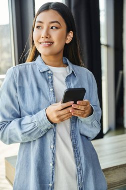 Güzel ve hırslı Asyalı iş kadını cep telefonu tutuyor, gülümsüyor ve pencerenin kenarına bakıyor çağdaş ofiste, mavi kot gömlek, esmer saçlar, parlak bir gülümseme