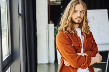 serious and fashionable bearded entrepreneur with long hair wearing trendy shirt, standing with crossed arms and looking at camera in contemporary office, hipster style, modern businessman clipart