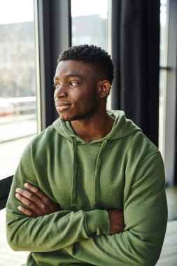 Düşünceli Afro-Amerikan iş adamı, yeşil kapüşonlu, kollarını kavuşturmuş, pencerenin kenarına bakan genç girişimci, 