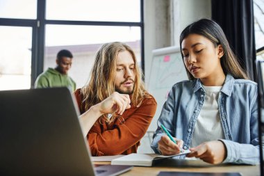 young asian woman writing in notebook near attentive long haired man and laptop on work desk in modern office, working on startup planning, partnership and collaboration clipart