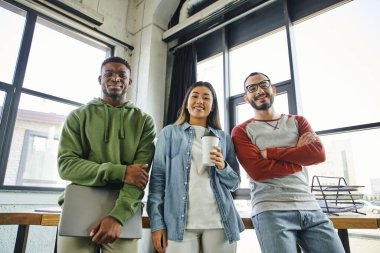 young and successful multiethnic business colleagues smiling at camera in modern office, african american man with laptop, asian woman with takeaway drink, bearded man in eyeglasses  clipart