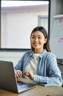 Mutlu Asyalı iş kadını, esmer saçlı, parlak gülüşlü, elinde kalemle iş yerinde bilgisayarının yanında otururken kameraya bakan. 