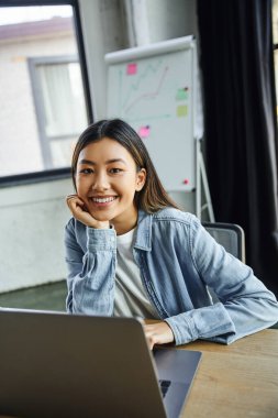 young and pleased asian businesswoman with brunette hair, in blue denim shirt looking at camera near laptop and flip chart with graphs on blurred background in contemporary office clipart