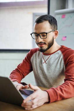 pozitif ve konsantre sakallı girişimci gözlüklü ve günlük kıyafetlerle iş yerinde oturuyor ve bilgisayarla modern ofiste bulanık bir ön planda çalışıyor başarılı bir genç