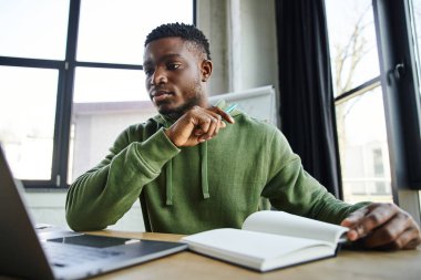 young and handsome african american businessman in green stylish hoodie looking at laptop near notebook while sitting at workplace and working on blurred foreground in modern office clipart