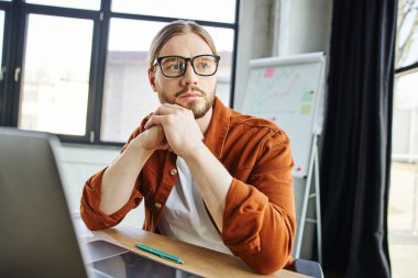 stylish and pensive businessman in eyeglasses and trendy casual clothes looking away near laptop and pen on work desk next to flip chart with graphs on blurred background in modern office clipart