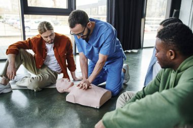 Profesyonel sağlık çalışanı kalp masajı mankeni üzerinde göğüs masajı yapıyor ve eğitim odasındaki çok ırklı genç katılımcılara kardiyopulmoner resüsitasyon gösteriyor