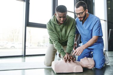 medical instructor in uniform and eyeglasses pointing at CPR manikin while african american man doing chest compressions, effective life-saving skills and emergency preparedness concept clipart