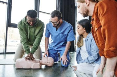 Genç çok ırklı insanlar ve tıbbi eğitmen, kalp masajı manikürü, kardiyopulmoner resüsitasyon ve ilk yardım eğitim semineri için göğüs kompresyonu yapan Afrikalı Amerikalıya bakıyor.