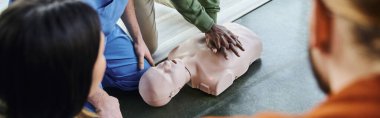 partial view of african american man doing chest compressions on CPR manikin near medical instructor and participants on blurred foreground, life-saving skills and techniques concept, banner clipart
