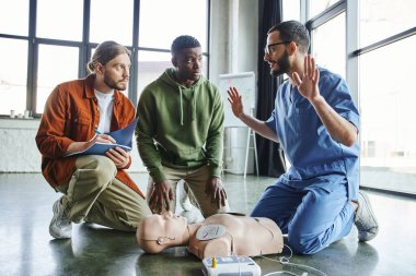healthcare worker gesturing and talking next to CPR manikin with defibrillator near african american participant and young man writing in notebook, effective life-saving skills and techniques concept clipart