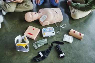 cropped view of multiethnic participants of first aid seminar near CPR manikin, defibrillator, wound care simulators, bandages, compression tourniquets and neck brace in training room, top view clipart