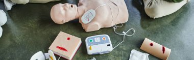 top view of CPR manikin, automated external defibrillator and wound care simulators near cropped participants of first aid training seminar, health care and emergency preparedness concept, banner clipart