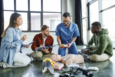 medical instructor showing wound care simulator to multiethnic team with clipboards near CPR manikin and medical equipment in training room, effective life-saving skills concept clipart
