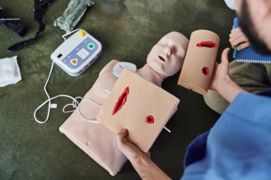 first aid training seminar, cropped view of healthcare worker holding wound care simulators near CPR manikin and automated external defibrillator, safety and emergency preparedness concept, top view clipart