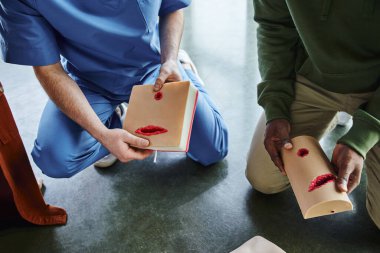 partial view of african american man and professional medical instructor holding wound care simulators during hands-on learning in training room, safety and emergency preparedness concept clipart