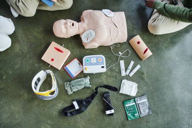first aid seminar, cropped view of young people near CPR manikin, defibrillator, wound care simulators, bandages, compression tourniquets, syringes and neck brace in training room, top view clipart