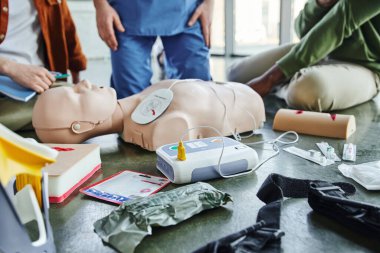 CPR manikin, defibrillator, wound care simulators, compression tourniquet, bandages and syringes near instructor and multiethnic participants of first aid seminar, emergency preparedness concept clipart