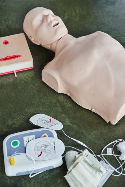 high angle view of CPR manikin near wound care simulator and automated external defibrillator on floor in training room, medical equipment for first aid training and skills development clipart