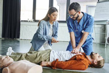 Tıp semineri, üniformalı sağlık çalışanı ve gözlüklü Asyalı kadına kalp masajı tekniklerini sunan kalp masajı manikinin yakınındaki eğitim odasında yatan adam, hayat kurtaran beceriler konsepti.