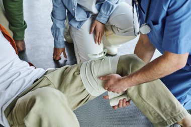 İlk yardım semineri sırasında çoklu etnik grupların yakınındaki bir adamın bacağına baskı uygulayan uzman sağlık görevlisinin kısmi görüşü, kanama önleme teknikleri kavramı