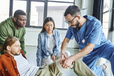 Profesyonel sağlık çalışanı, eğitim odasındaki ilk yardım semineri sırasında çok ırklı öğrencilerin yakınındaki bir adamın bacağına bandaj yapıştırıyor, acil durum hazırlık konseptinin önemi