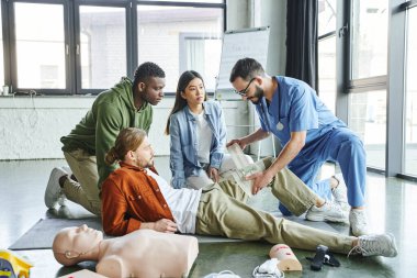 medical training, professional instructor in uniform and eyeglasses bandaging leg of man near multicultural participants, CPR manikin and medical equipment, emergency preparedness concept clipart