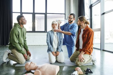 professional paramedic in eyeglasses and uniform bandaging head of asian woman near interracial men during medical seminal with CPR manikin and medical equipment, effective first aid concept clipart
