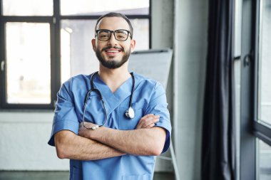 Gözlüklü ve mavi üniformalı neşeli ve sakallı doktor kameraya, ilk yardım seminerine ve acil durum hazırlık konseptine bakarken kolları bağlı ve steteskoplu duruyor.